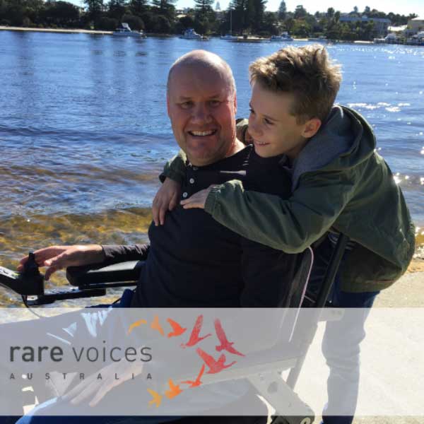 Photo of Damian in a wheelchair with his son standing behind and hugging him